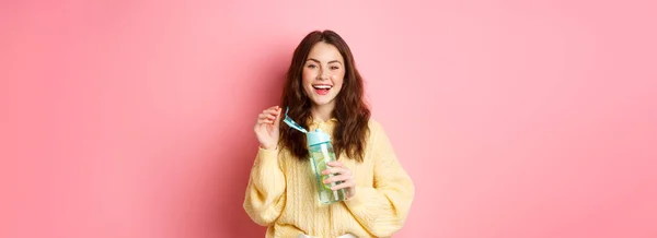 stock image Portrait of happy brunette woman with curly hairdo, drinking water with lemon from personal bottle, laughing and smiling, feeling healthy, standing against pink background.