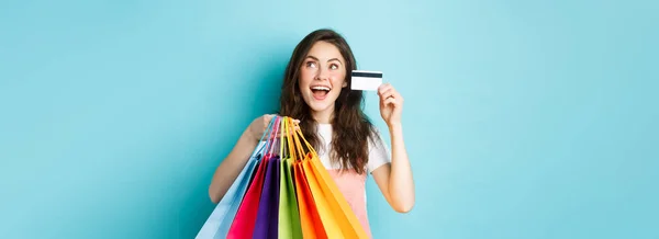 快乐的黑发女孩 带着塑料信用卡和购物袋的购物狂 站在蓝色的背景上 看着商店的标志或横幅 看起来像在做梦 — 图库照片