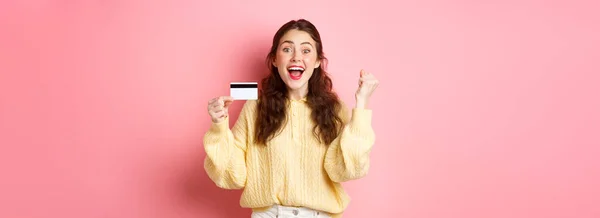 兴奋的年轻女子获奖 看到令人敬畏的促销 展示她的塑料信用卡和欢乐的尖叫 说是的 站在粉红的背景下 — 图库照片