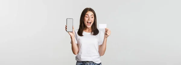 スマートフォンの画面とクレジットカード 白い背景を示す興奮した魅力的な女の子 — ストック写真