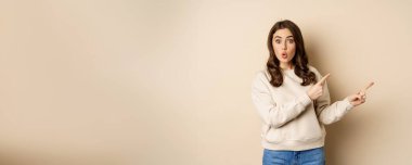 Surprised brunette woman pointing fingers right, showing advertisement and say wow impressed, standing over beige background.