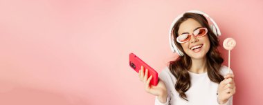 Close up portrait of stylish glamour girl, beautiful woman in headphones, listening music with smartphone, wearing sunglasses and licking lolipop.