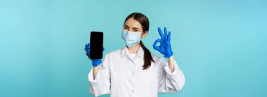 Portrait of doctor in medical face mask and gloves, showing mobile phone app, smartphone screen and okay sign, recommending online checkup website, standing over torquoise background.