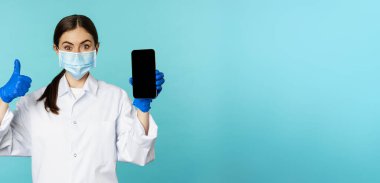 Portrait of doctor in medical face mask and gloves, showing mobile phone app, smartphone screen and thumb up, recommending online checkup website, standing over blue background.