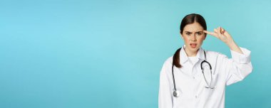 Annoyed woman doctor pointing finger at head, scolding someone stupid, crazy or strange, standing over torquoise background.