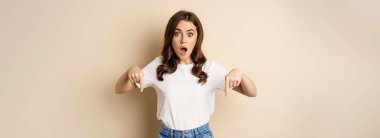 Surprised modern womn pointing fingers down, gasping amazed, showing smth awesome below, standing over beige background.