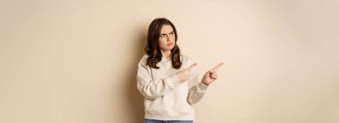 Woman pointing left and staring confused at banner copy space, strange logo, standing over beige background.