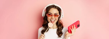 Close up portrait of stylish glamour girl, beautiful woman in headphones, listening music with smartphone, wearing sunglasses and licking lolipop.