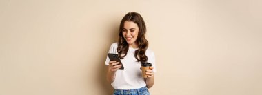 Smiling woman using mobile phone app and credit card, concept of online shopping, contactless payment and delivery, standing over beige background.