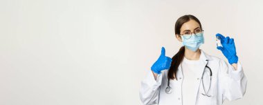 Profesyonel doktor, tıbbi maske ve gözlüklü doktor, aşıyı gösteren ve onaylayan, covid-19 'dan aşı olmayı öneren genç kadın portresi..
