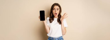 Surprised woman gasping, showing smartphone app, mobile screen and looking amazed, standing in casual clothes against beige background.