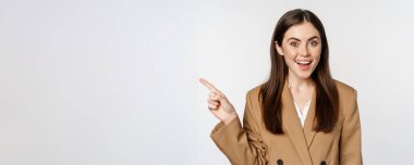 Portrait of businesswoman pointing finger left, showing corporate banner, logo, standing in brown suit over white background.