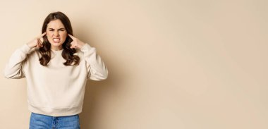 Annoyed and angry woman shut ears, complaining at loud noisy music noise, standing over beige background.