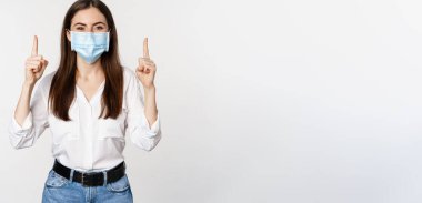 Portrait of corporate woman in face medical mask from coronavirus, pointing fingers up, showing advertisement, white background.