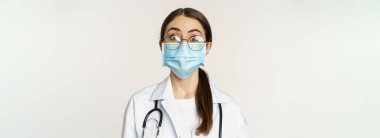 Medical worker, young doctor looking amazed and surprised, wearing face mask to prevent catching covid-19, standing over white background.