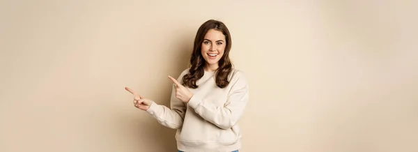 stock image Smiling gorgeous woman pointing fingers right, inviting people, showing logo or banner, standing over beige background.