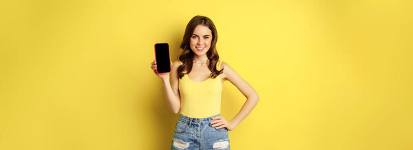 Stylish smiling woman showing mobile phone screen, app interface on smartphone, standing in tank top and shorts against yellow background.