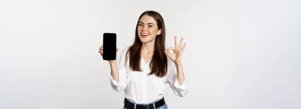 満足した顔をした幸せな笑顔の女性 優れたアプリをお勧めします 携帯電話の画面を表示 大丈夫なサイン 白い背景の上に立って — ストック写真
