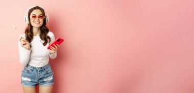 Stylish brunette woman dancing in headphones with mobile phone, listening music in earphones, licking lolipop, standing over pink background.