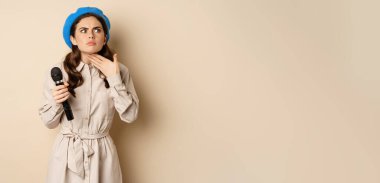 Stylish singer, woman with microphone touching throat, feeling ill, standing over beige background. Copy space