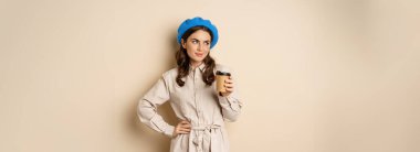 Beautiful happy girl drinking takeaway coffee from cafe and smiling, posing with cup of beverage, beige background.