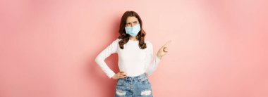 Confused, indecisive young woman in medical face mask, pointing finger left and looking complicated, making decision, standing over pink background.