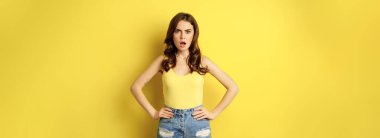Shocked stylish girl hold hands on waist, drop jaw and stare confused at camera, posing in summer clothes over yellow background.