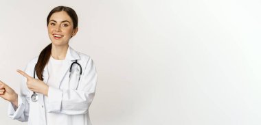 Portrait of smiling young woman doctor, healthcare medical worker, pointing fingers left, showing clinic promo, logo or banner, standing over white background.