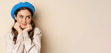 Close up portrait of feminine young woman in trendy french hat, posing cute and romantic, making silly coquettish face and tender smile, beige background.