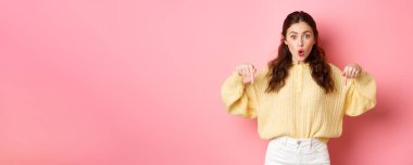 Vay canına, süper havalı. Heyecanlı esmer kız parmaklarını aşağı işaret ediyor, nefes nefese merak ediyor, yön gösteriyor, alt logoda işaret ediyor, pembe arka planda duruyor..