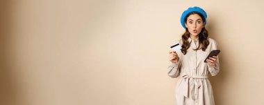 Portrait of girl looking excited, say wow, holding credit card and mobile phone, standing over beige background. Copy space