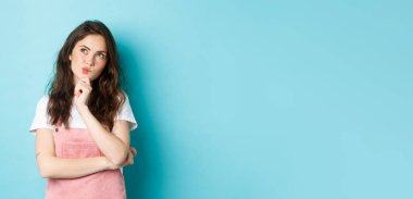 Düşünceli esmer kadın seçim yapıyor, çene ve düşünerek el ele tutuşuyor, düşünceli bir yüzle sol üst köşeye bakıyor, mavi arka planda duran bir şeyi düşünün..