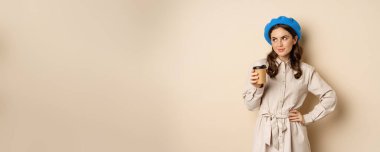 Beautiful happy girl drinking takeaway coffee from cafe and smiling, posing with cup of beverage, beige background.