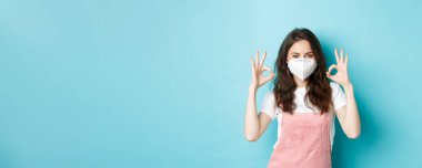 Covid, sağlık ve salgın hastalık konsepti. Solunum cihazında tatmin olmuş güzel bir kız, tıbbi maske onaylama işareti, koronavirüsten önleyici önlemler, mavi arka plan..