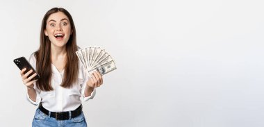 Online microcredit loans and banking concept. Happy woman holding mobile phone and money, smiling and laughing, standing over white background.