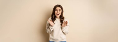 Image of surprised, amazed brunette girl gasping, looking fascinated at camera, standing over beige background.