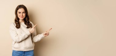 Smiling gorgeous woman pointing fingers right, inviting people, showing logo or banner, standing over beige background.