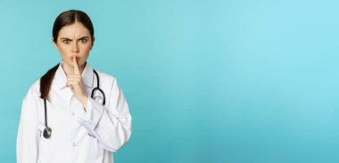 Angry female doctor, healthcare medical worker shushing with disapproval, taboo quiet gesture, silence someone, standing over torquoise background.