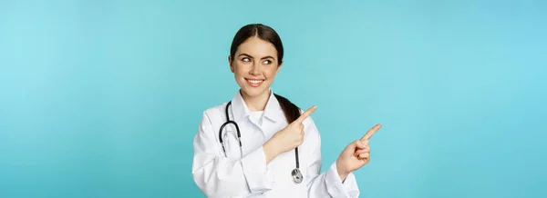 笑顔の医療従事者の肖像 聴診器付き白衣の少女医師 指差し指左 医療クリニックの広告を表示 ターコイズの背景 — ストック写真