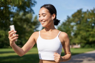 Su şişesi ve akıllı telefonlu fitness kadını, parkta koşuyor ve gülümsüyor, cep telefonu uygulamasına bakıyor, spor uygulamalarını kontrol ediyor..