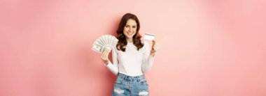 Credit, money and loans concept. Happy beautiful girl holding credit card and cash, looking satisfied, thinking of shopping, standing over pink background.