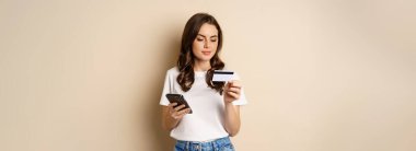Image of young happy woman paying online, holding smartphone and credit card, enter info in application on mobile phone, standing against beige background.