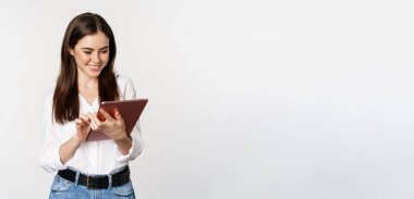 Portrait of smiling corporate woman looking at digital tablet, working, standing over white background.