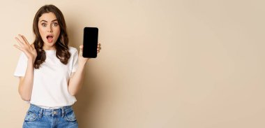 Surprised woman gasping, showing smartphone app, mobile screen and looking amazed, standing in casual clothes against beige background.
