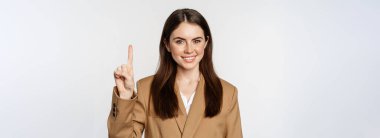 Portrait of corporate woman, saleswoman showing number one finger and smiling, standing in suit over white background.