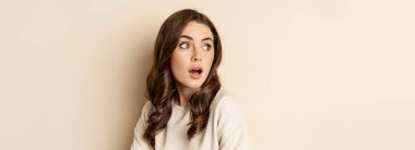Close up portrait of brunette feminine woman turn back, look behind with surprised face expression, standing against beige background.