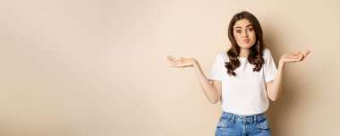Young confused woman shrugging shoulders and with clueless face expression, standing over beige background.