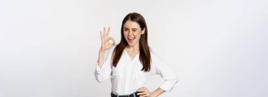 Smiling confident business woman, winking, showing okay ok sign, excellent, no problem gesture, standing over white background.