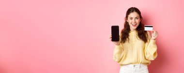 Teknoloji ve online alışveriş. Plastik kredi kartı ile boş akıllı telefon ekranı gösteren genç ve çekici bir kadın model. Banka hesabı ya da başvuru formunu pembe arka planda duruyor..