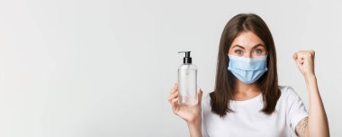 Covid-19, sağlık ve sosyal uzaklık kavramı. Tıbbi maskeli, el dezenfektanı gösteren çekici kızın yakın çekimi, antiseptik öneriyor..